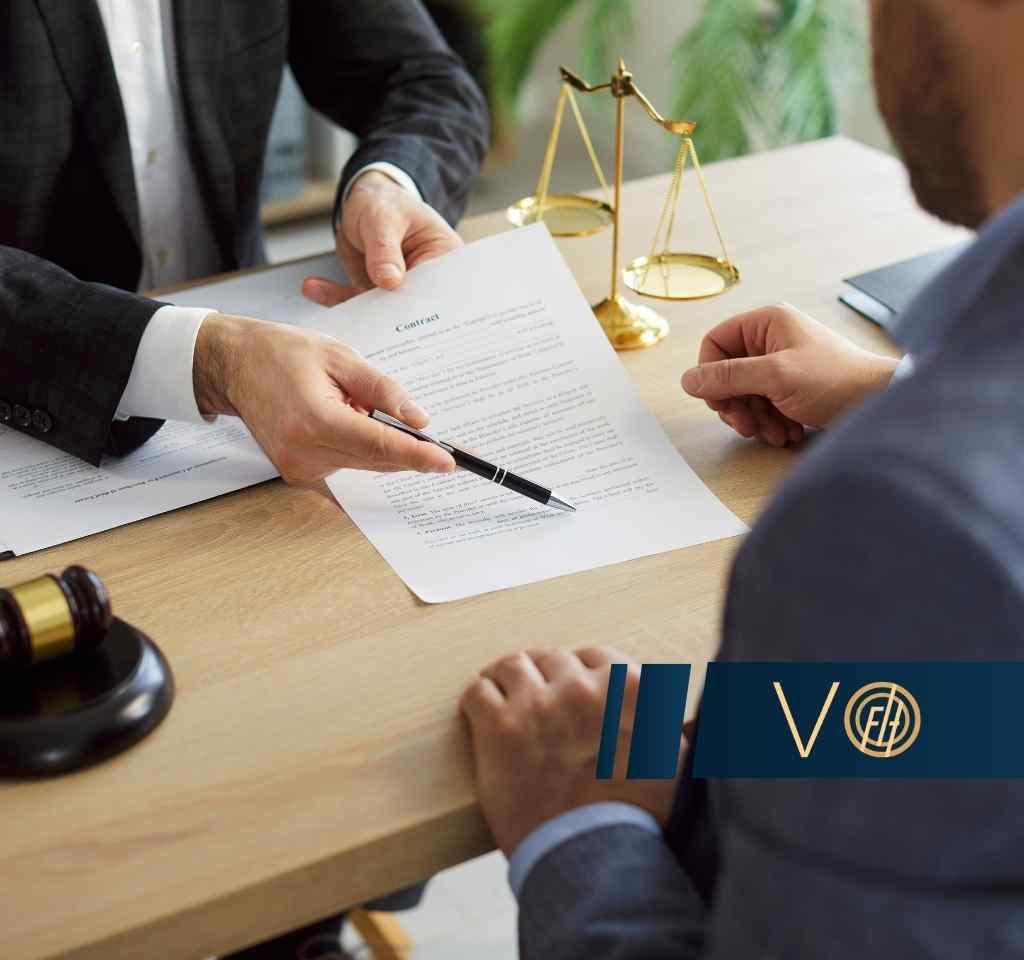 Na drewnianym stole leży dokument prawny, na który dłoń w garniturze wskazuje długopisem, obok leżą młotek sędziowski i waga szalkowa