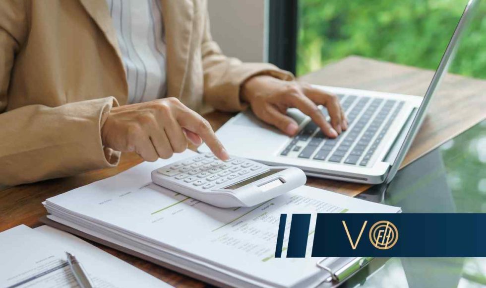 Kobieta w beżowej marynarce siedzi przy biurku z laptopem, używając kalkulatora na stosie dokumentów