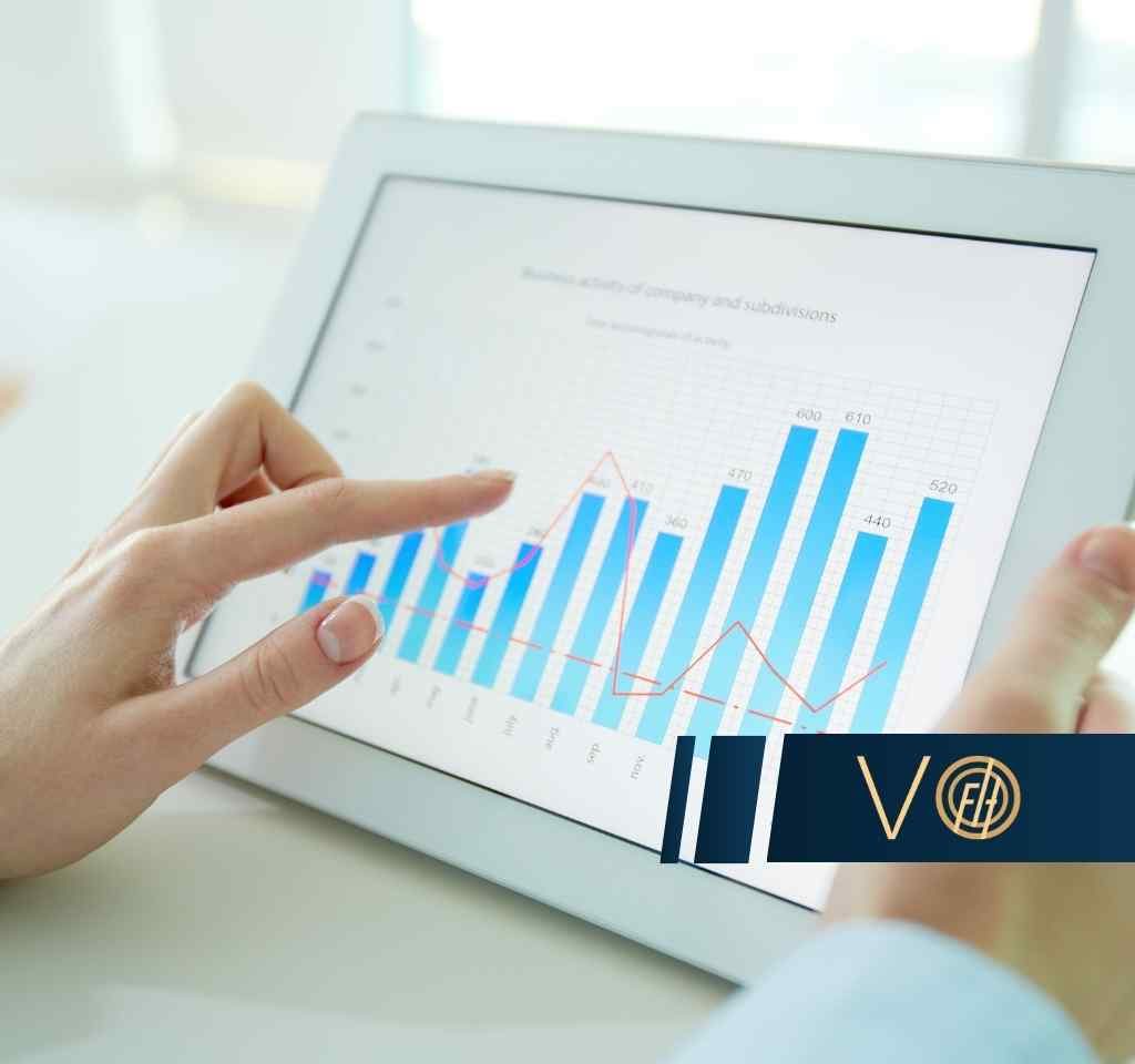 Dłonie trzymają tablet, na którego ekranie wyświetlony jest niebieski wykres słupkowy z rosnącą czerwoną linią, a palec wskazuje na dane na wykresie