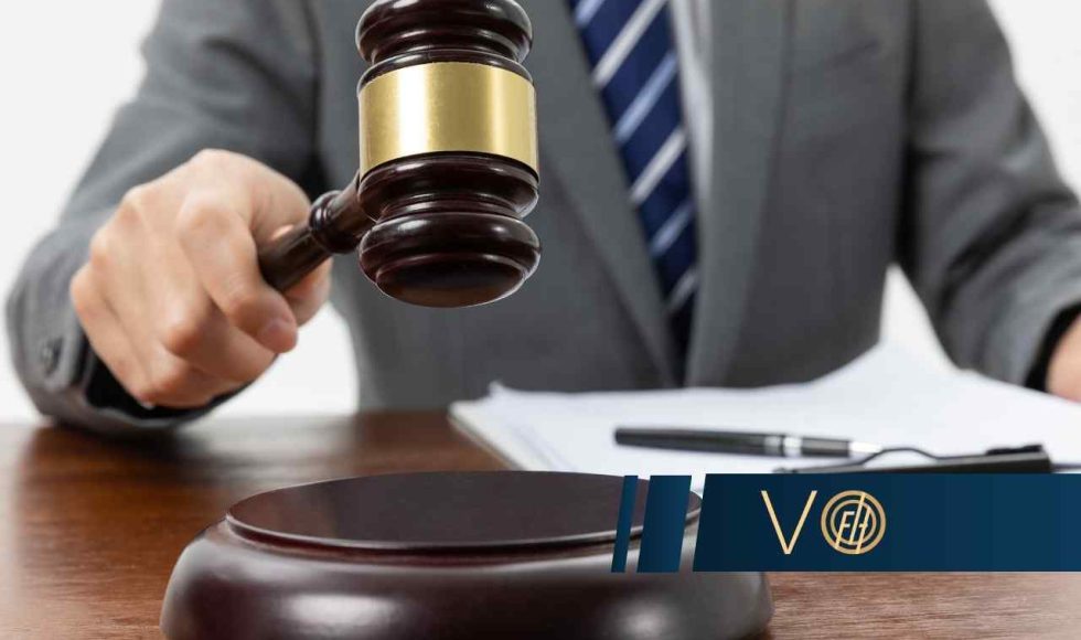 Dłoń w mankiecie garnituru trzyma młotek sędziowski nad jego podstawką, leżącymi na drewnianym biurku, na tle dokumentów