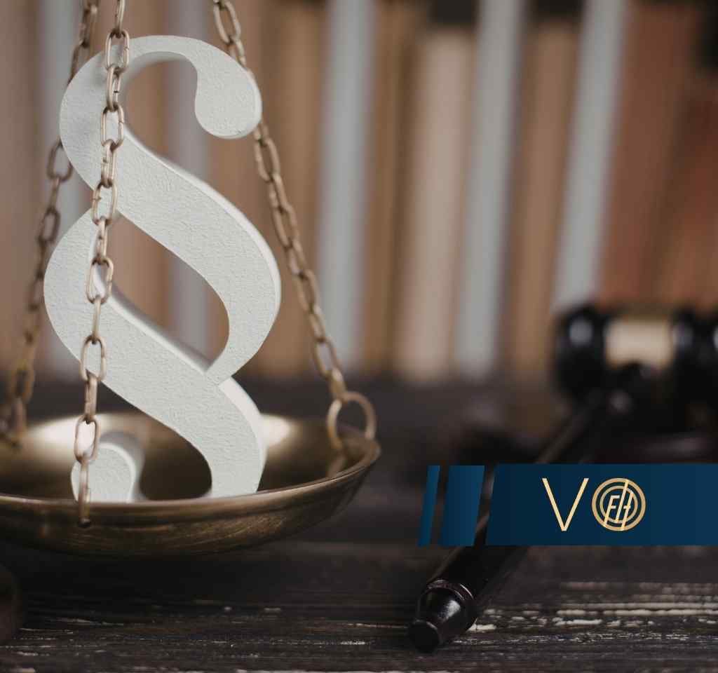 Na metalowej szali wagi szalkowej, zawieszonej na łańcuchach, leży duży, biały symbol paragrafu, a w prawym dolnym rogu widoczny jest fragment młotka sędziowskiego