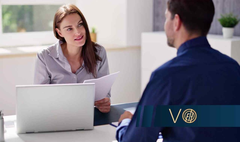 Kobieta w jasnej koszuli siedzi za biurkiem z laptopem i dokumentami, rozmawiając z mężczyzną siedzącym tyłem do obiektywu