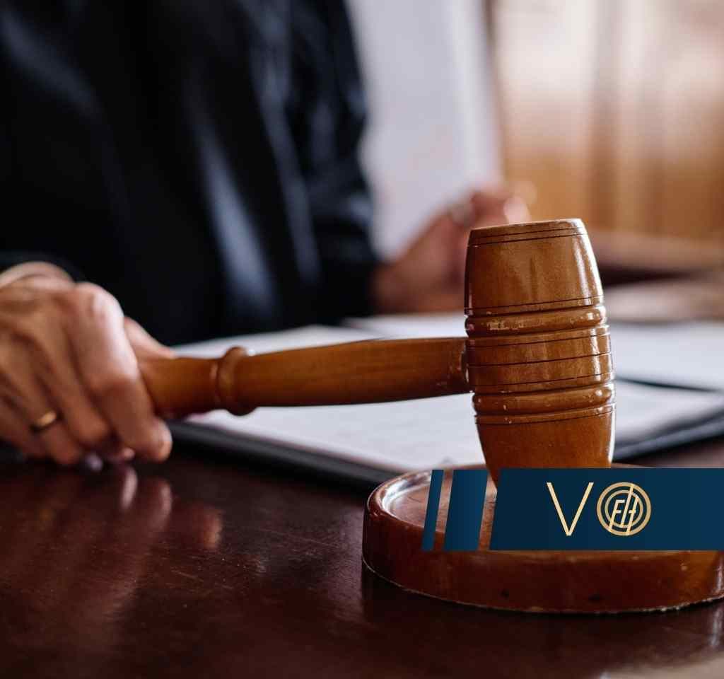 Zbliżenie na dłoń sędziego trzymającą drewniany młotek oparty o podstawkę na biurku z dokumentami