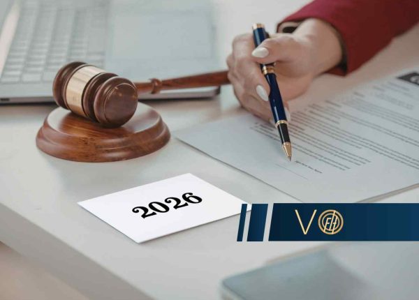 Osoba trzyma eleganckie pióro nad dokumentem obok sędziowskiego młotka i kartki z napisem „2026”.