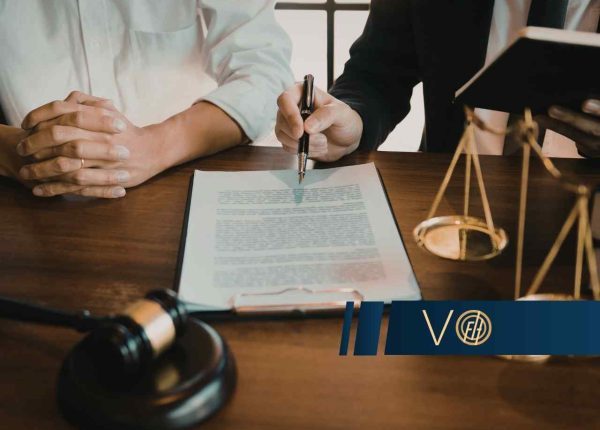 Zbliżenie na dłonie osób podpisujących dokument przy biurku, na którym stoją atrybuty prawnicze: waga sprawiedliwości oraz młotek sędziowski.