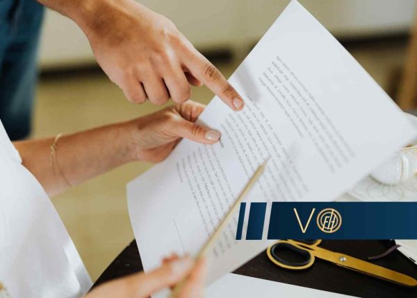 Dwie osoby analizują dokument papierowy; jedna trzyma pismo, a druga wskazuje palcem na konkretny akapit tekstu. Na biurku obok widoczne są nożyczki oraz eleganckie logo w ciemnym kolorze.