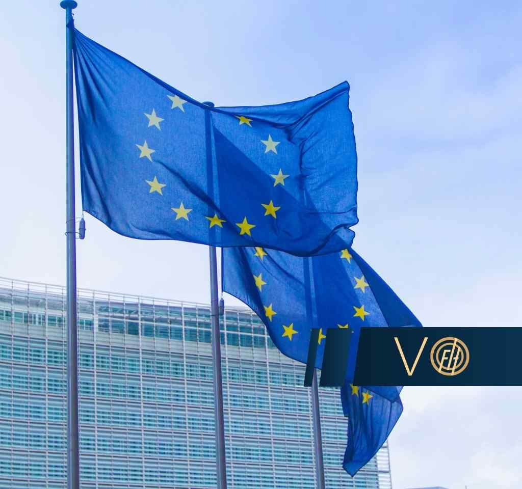 Kilka flag Unii Europejskiej z żółtymi gwiazdami na niebieskim tle powiewa na masztach przed nowoczesnym, przeszklonym budynkiem instytucji unijnej, na tle jasnego nieba.