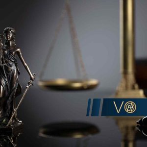 Ciemnobrązowa statuetka Temidy z mieczem i wagą stoi na błyszczącym blacie, z rozmytym złotym elementem wagi w tle.