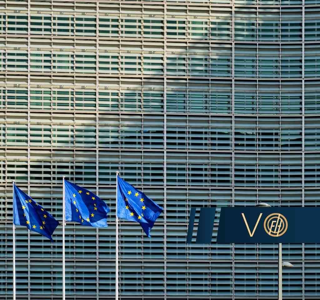European Union flags on flagpoles in front of a modern glass-front building with a dark blue banner displaying a logo