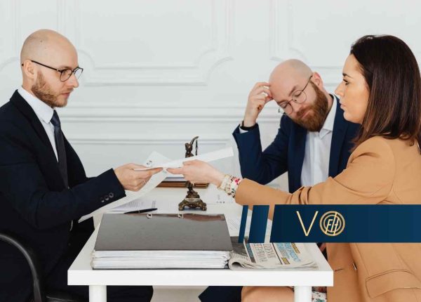 Scena w biurze lub kancelarii; po lewej stronie mężczyzna w ciemnym garniturze podaje dokument kobiecie w beżowej marynarce. W tle widać drugiego mężczyznę, który z przejęciem trzyma się za głowę. Na pierwszym planie leżą teczki z dokumentami i gazeta.