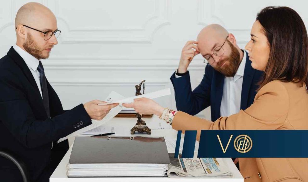 Scena w biurze lub kancelarii; po lewej stronie mężczyzna w ciemnym garniturze podaje dokument kobiecie w beżowej marynarce. W tle widać drugiego mężczyznę, który z przejęciem trzyma się za głowę. Na pierwszym planie leżą teczki z dokumentami i gazeta.