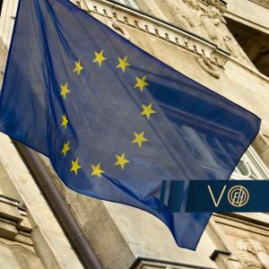 Flaga Unii Europejskiej powiewająca na maszcie przymocowanym do elewacji zabytkowego budynku z ozdobnymi gzymsami i detalami architektonicznymi.