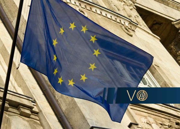 Flaga Unii Europejskiej powiewająca na maszcie przymocowanym do elewacji zabytkowego budynku z ozdobnymi gzymsami i detalami architektonicznymi.