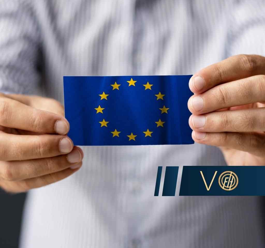 Zbliżenie na dłonie osoby w jasnej, prążkowanej koszuli, która trzyma małą kartę z flagą Unii Europejskiej.