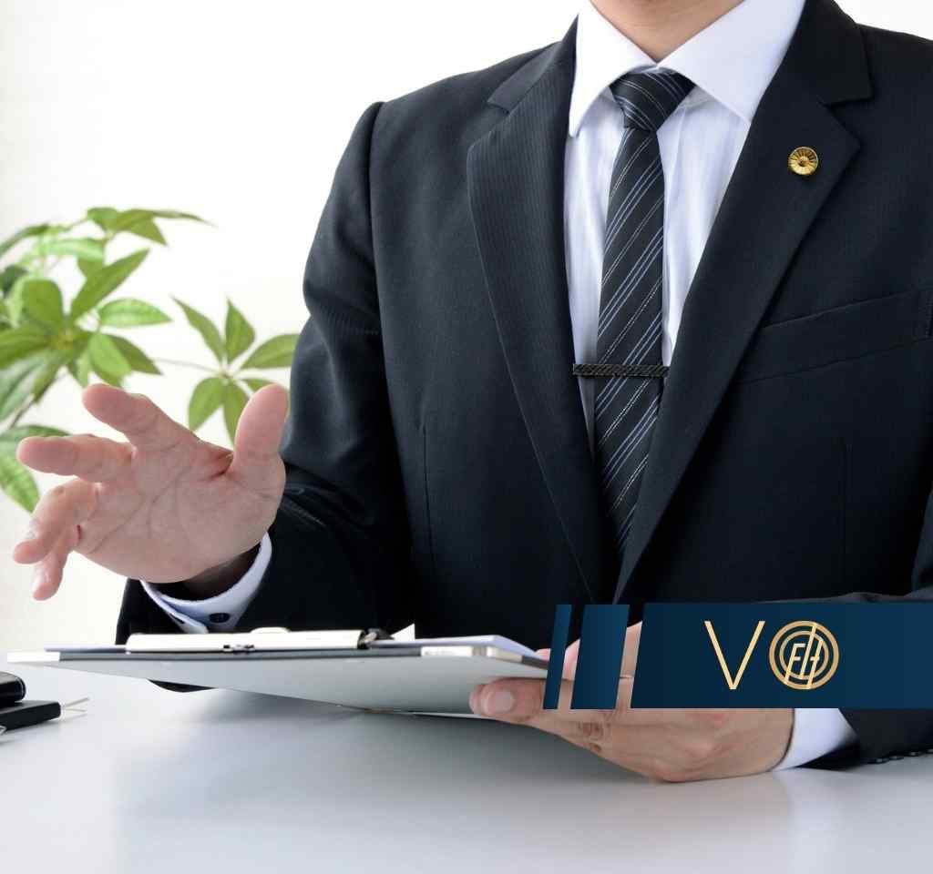 Businessperson in a dark suit gestures with an open hand at a desk, clipboard and pens nearby.