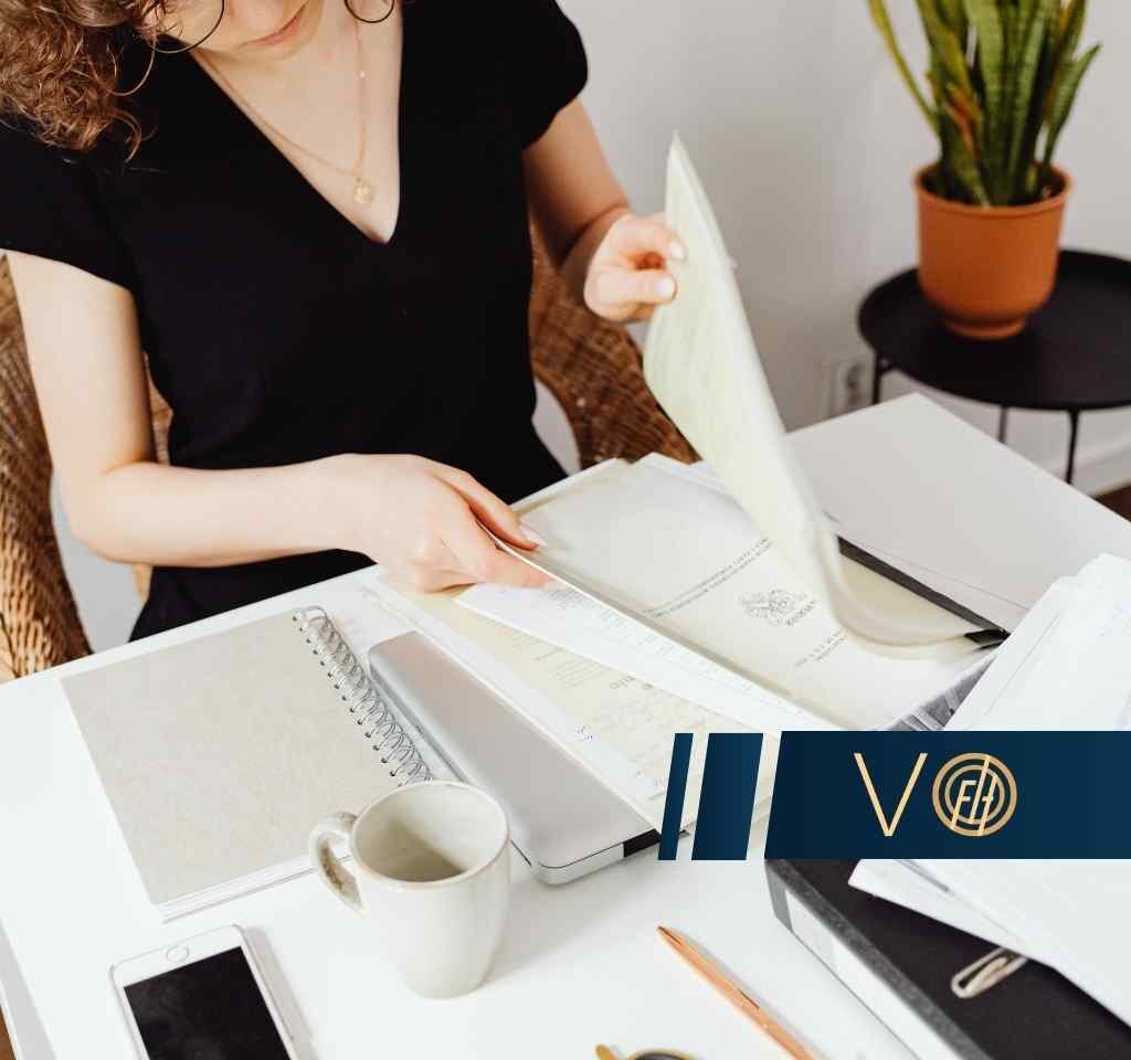 Woman in a black top flipping through documents at a white desk, with a notebook, mug, and plant nearby. Logo overlay in the bottom right.