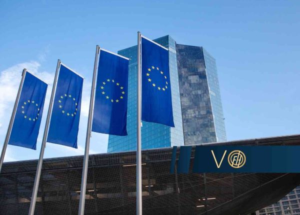 Four blue European Union flags with yellow stars on tall flagpoles in front of a modern glass office building against a clear blue sky.
