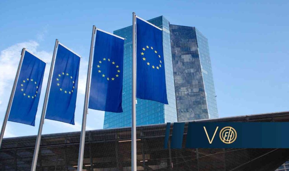 Four blue European Union flags with yellow stars on tall flagpoles in front of a modern glass office building against a clear blue sky.