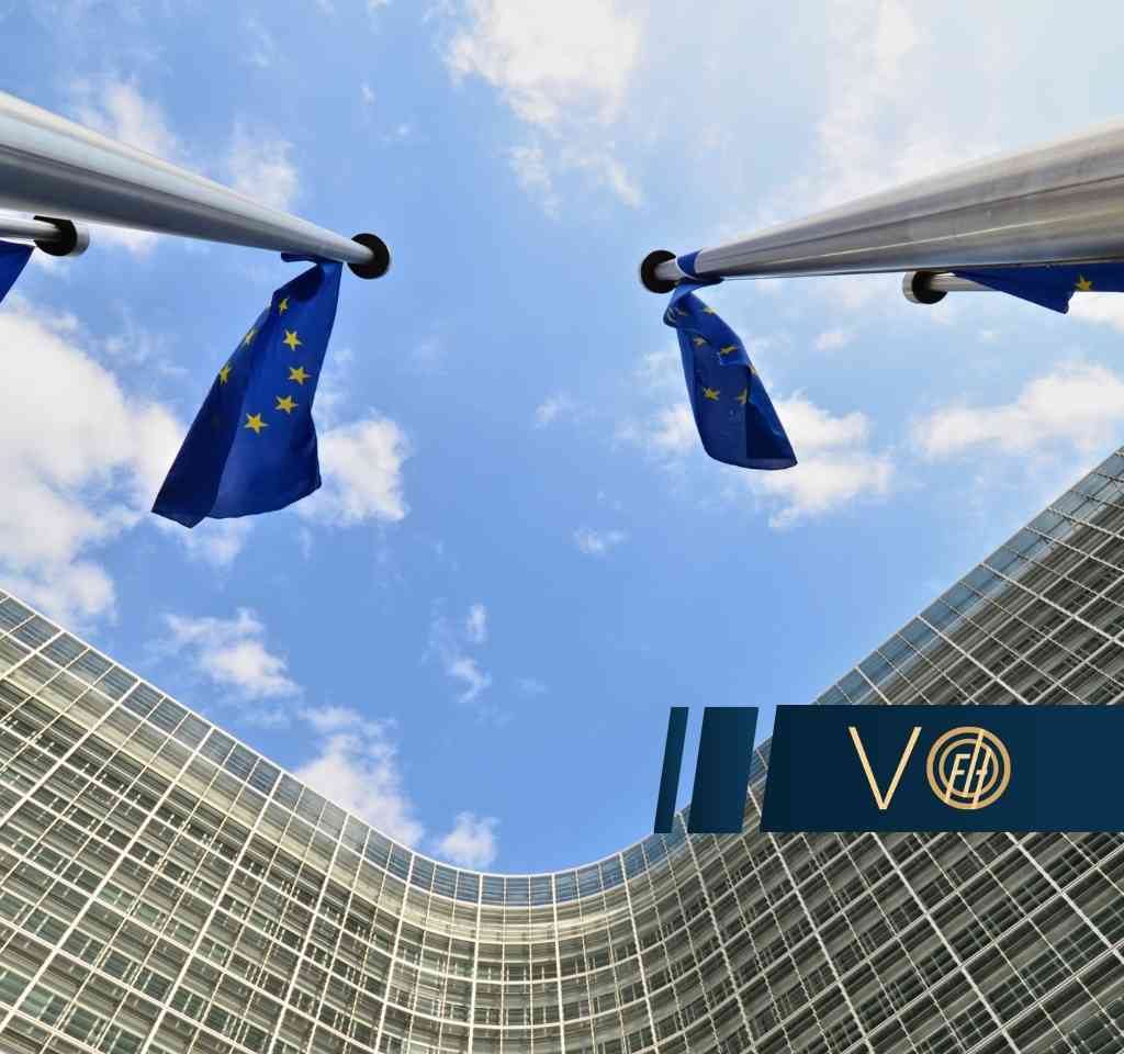 European Union flags flutter between flagpoles above a curved glass office building against a bright blue sky, political/ corporate setting