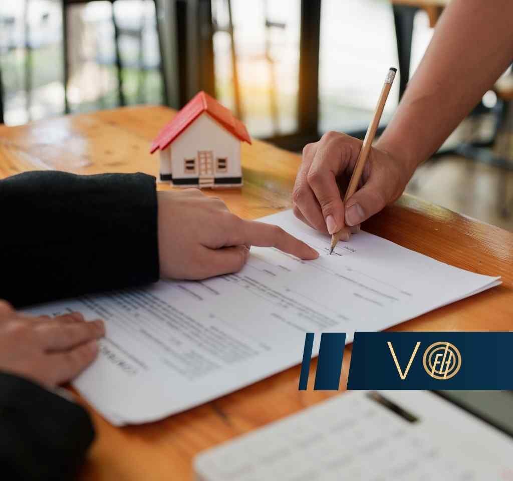 Person signs a document at a wooden table with a small house model nearby and a blue VO logo banner in the foreground (indicating real estate or contract signing).