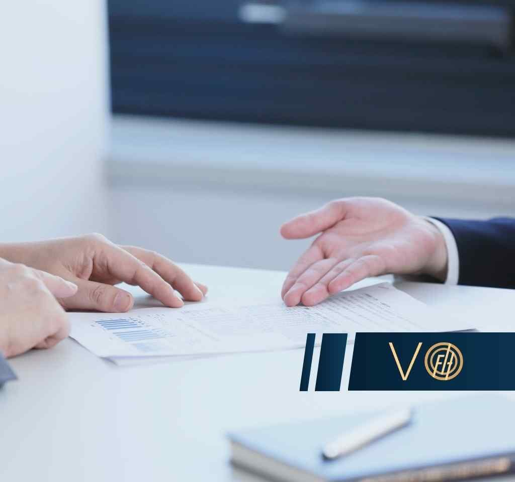 Two professionals review a document on a white table, with a dark blue branded banner on the lower right.