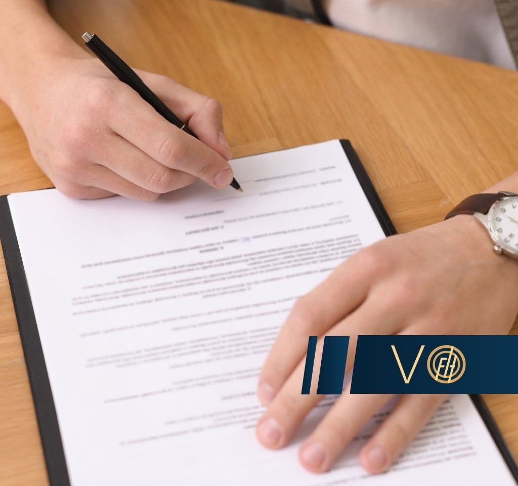 Person signing a document in a black-bound folder on a wooden table, with a silver watch visible on the wrist and a blue logo watermark at the bottom right.