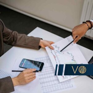 Two professionals review printed stock charts on a desk, with a smartphone and tablet nearby while a blue logo banner sits in the foreground.