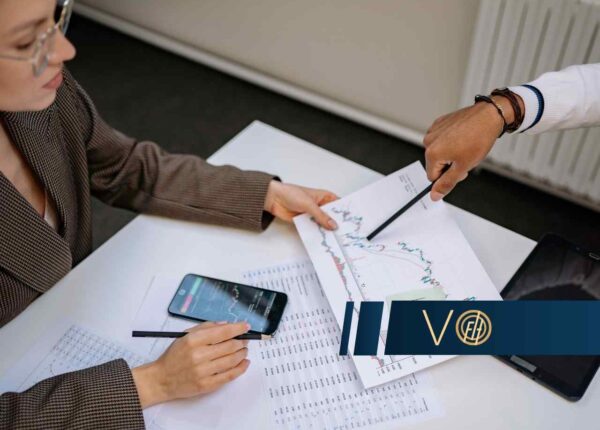 Two professionals review printed stock charts on a desk, with a smartphone and tablet nearby while a blue logo banner sits in the foreground.