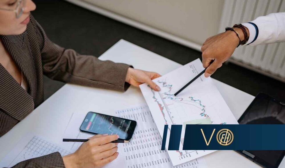 Two professionals review printed stock charts on a desk, with a smartphone and tablet nearby while a blue logo banner sits in the foreground.