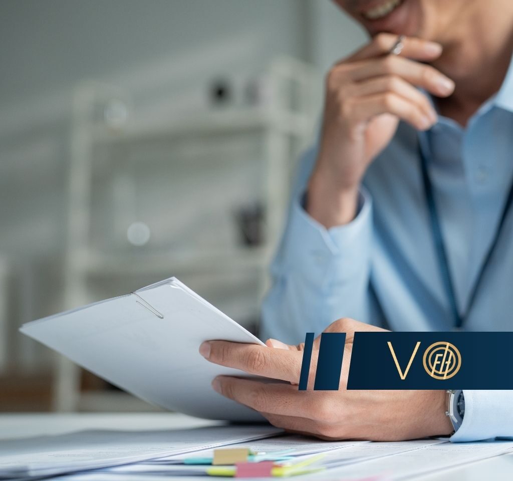Professional reviewing documents at a desk with sticky notes and papers; logo overlay on the right side of the image.