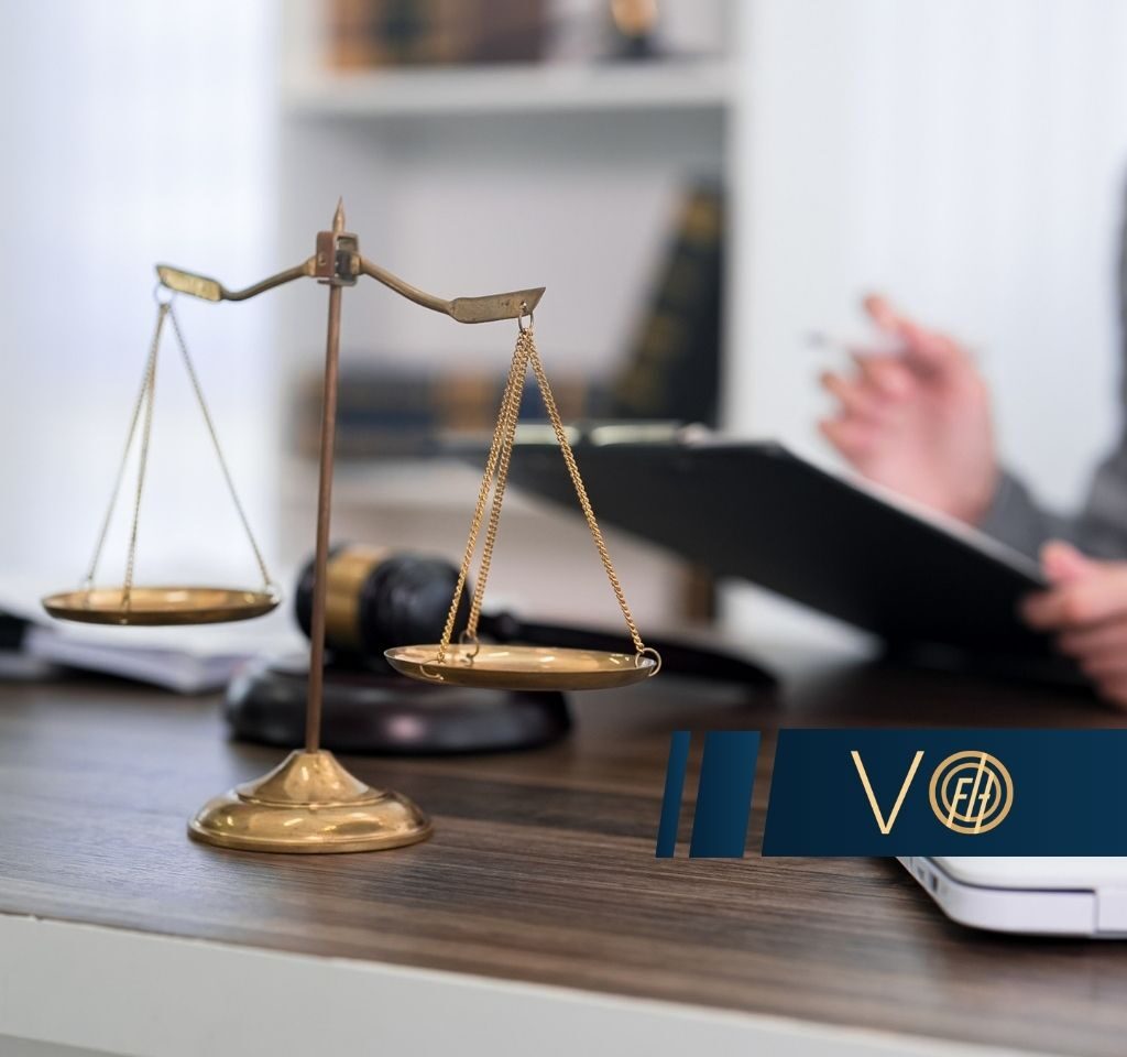 Brass balance scales on a wooden desk with a blurred lawyer reviewing documents in the background.
