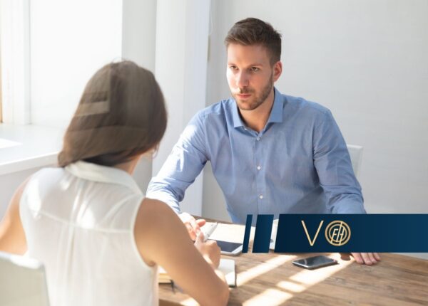 Man in a blue shirt sits across from a woman at a wooden table in a bright office, discussing documents in a business meeting.