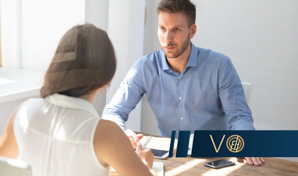 Man in a blue shirt sits across from a woman at a wooden table in a bright office, discussing documents in a business meeting.