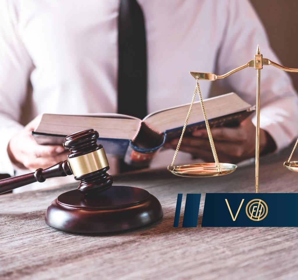 Gavel and scales of justice on a wooden desk, with a person reading a legal book in the background (lawful context).