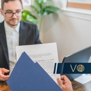 Professional setting: a person in a suit hands over a blue folder with papers across a wooden table, laptop nearby.
