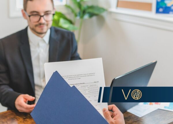 Professional setting: a person in a suit hands over a blue folder with papers across a wooden table, laptop nearby.