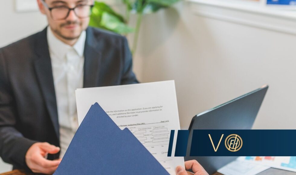 Professional setting: a person in a suit hands over a blue folder with papers across a wooden table, laptop nearby.
