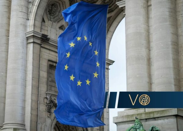 Blue European Union flag with 12 yellow stars draped between historic stone columns on a grand building, with a dark banner in the foreground.