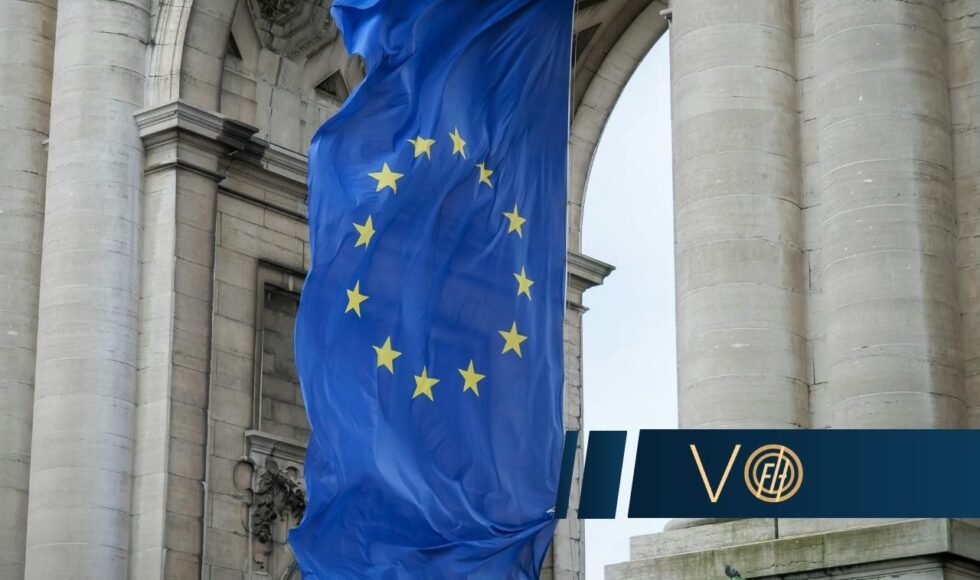 Blue European Union flag with 12 yellow stars draped between historic stone columns on a grand building, with a dark banner in the foreground.