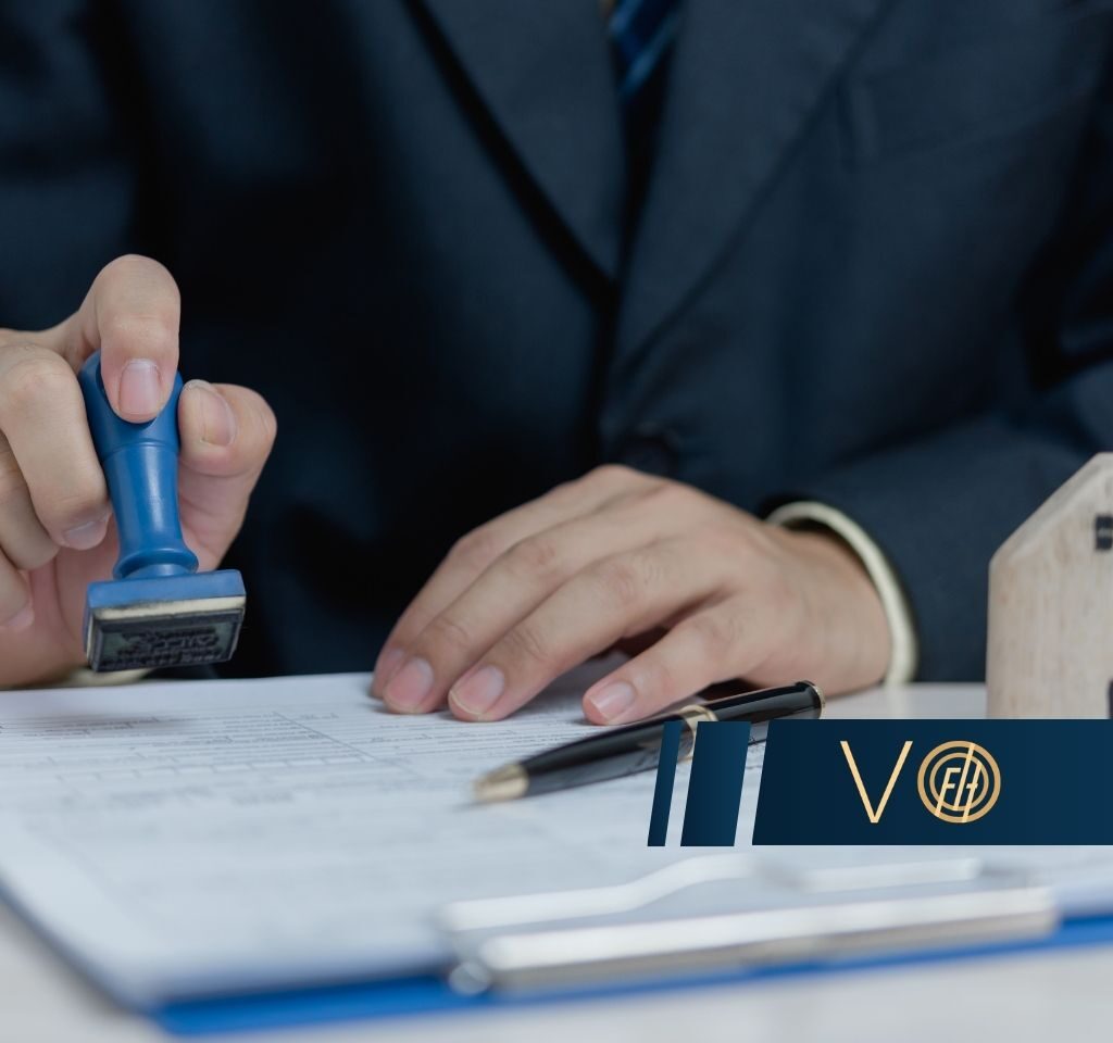 Close-up of a hand stamping a contract with a blue stamp, a pen nearby and a small wooden house model on the desk